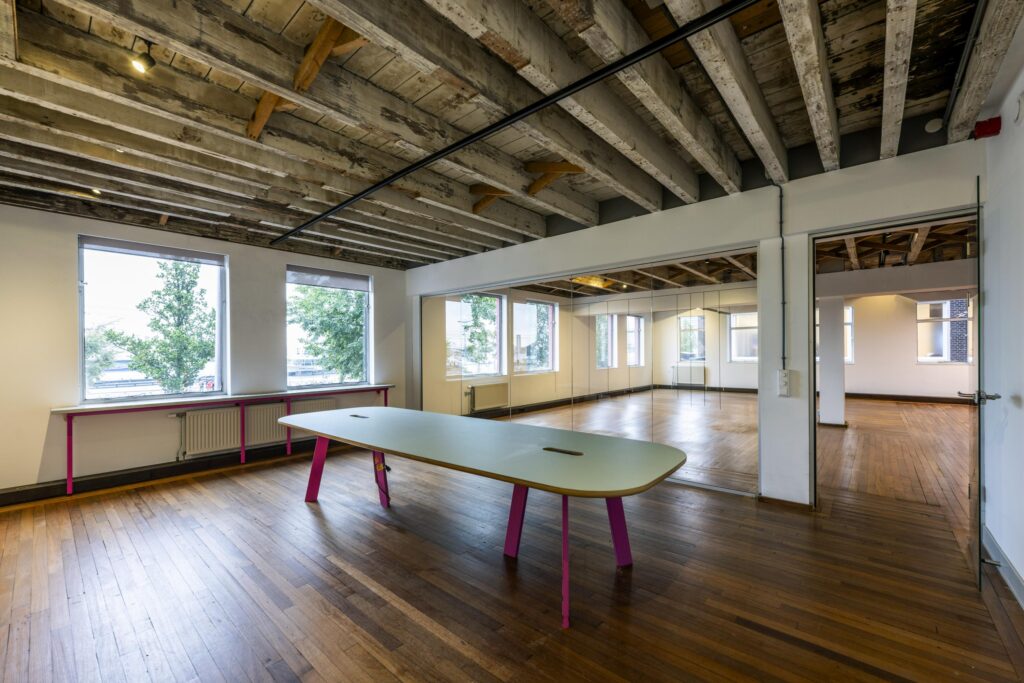 Licht en modern kantoorinterieur aan de De Ruijterkade met houten vloer, zichtbaar balkenplafond en opvallende tafel met roze poten.